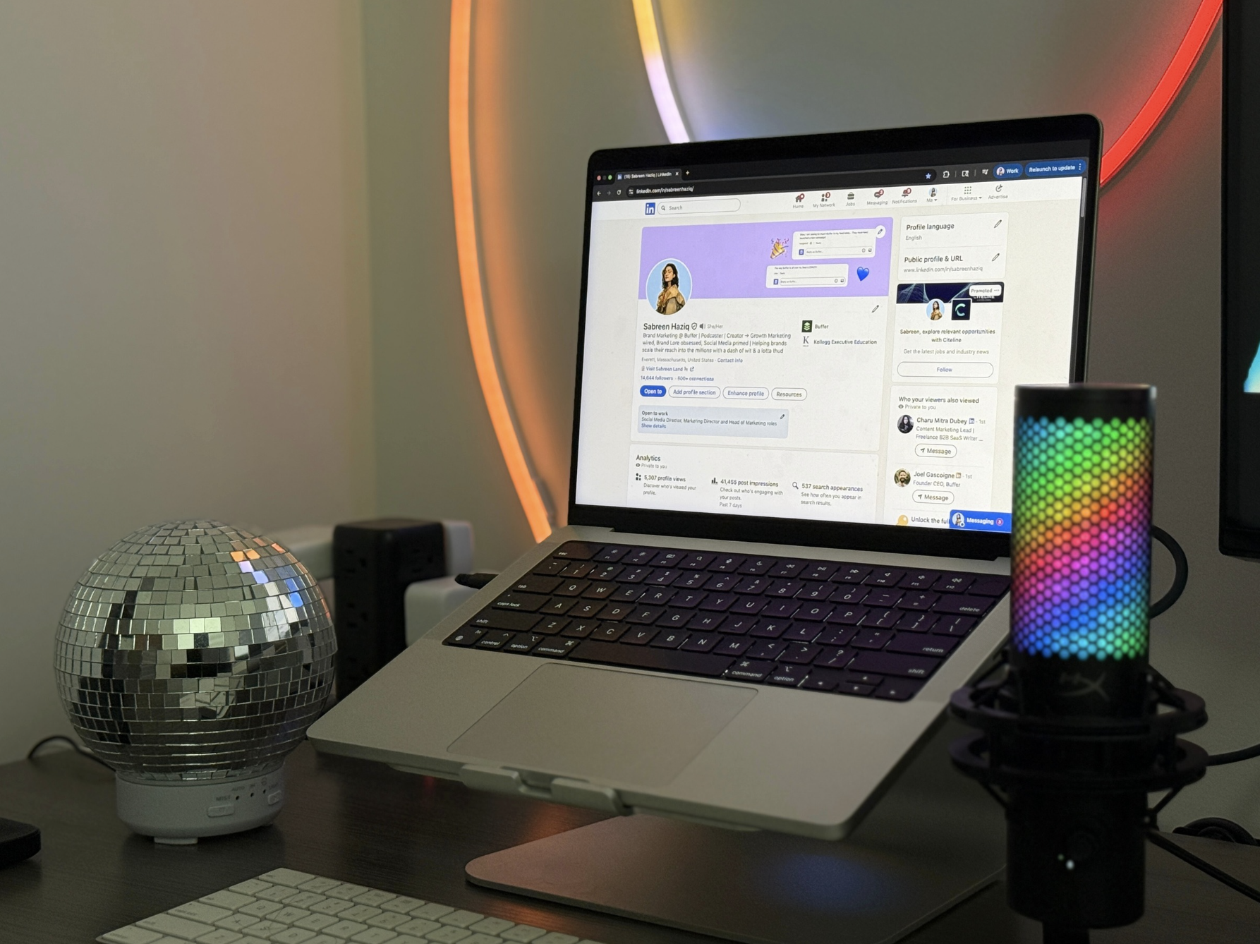 An open laptop on a desk showing Sabreen's LinkedIn profile, along with a multi-color LED mic and disco ball